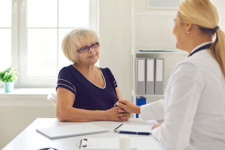 Young woman doctor touching senior positive woman patients hands during visit or consultation in medical clinic interior. Visiting doctor and modern healthcare support conceptの写真素材