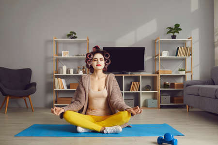 Finding moment of peace and time for self care in hectic domestic lifestyle. Happy calm relaxed woman in beauty face mask and curlers sitting in Lotus pose on yoga mat and meditating with eyes closedの写真素材