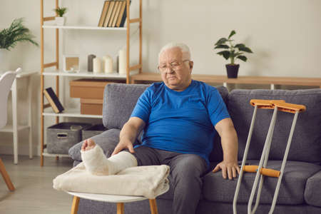 Rehabilitation after accident: Senior patient with bone fracture spends recovery period at home. Sad old man with broken leg in plaster cast sitting on sofa in living-room, thinking about his injuryの写真素材