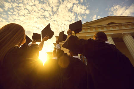 Group of young people university graduates in traditional masters mantles standing and holding diplomas in raised hands over sunset and university building background, bottom rear viewの写真素材