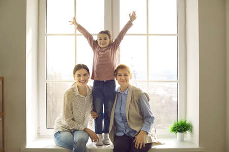 Portrait of happy multi generational family on windowsill at home. Cheerful mother, daughter and grandmother spending time together. Good multigenerational relationship, confidence in future conceptの写真素材