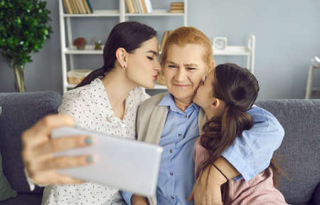 Happy grandmother, mother and grandkid capturing life moments they enjoy together on family holiday. Young daughter and little granddaughter kissing grandma on cheeks and taking selfie on mobile phoneの写真素材