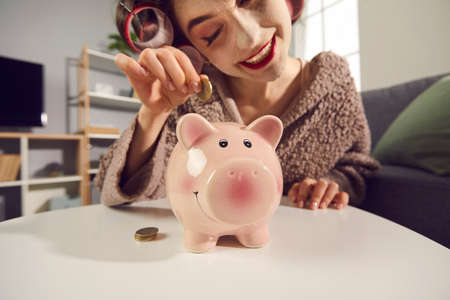 Happy young lady saving up some money. Funny wife, housewife or single woman making plans for the future and putting coins in her piggy bank. Concept of managing personal household budgetの写真素材