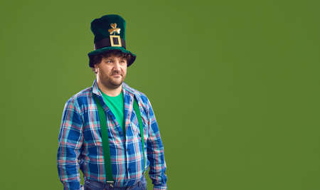 Serious pot-bellied caucasian bearded man in funny Leprechaun hat with gold with clover looking aside stands isolated on green copy space background. Saint Patrick Day party banner or cardの写真素材