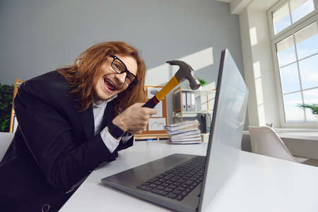 Funny insane office worker breaking down his computer. Crazy happy young man wants to smash laptop with hammer. Work stress, outdated software, deleting data, getting rid of broken equipment conceptの写真素材
