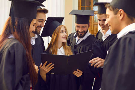 Group of young smiling happy students university graduates standing looking at girl mate diploma all together, university building at background. Graduation from university, education, diploma conceptの写真素材
