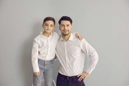 Positive father and son in stylish white shirts and jeans standing hugging and looking at camera over grey wall background. Happy fatherhood, childhood, family conceptの写真素材