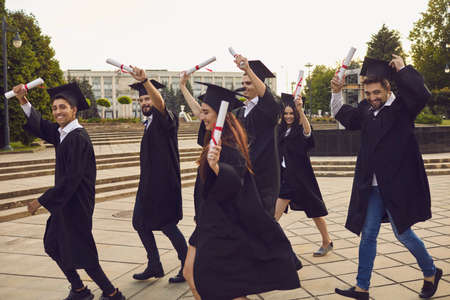 Group of happy young people university graduates in traditional bonets and masters mantles walking with diplomas in raised hands and looking at camera over university building background, side viewの写真素材