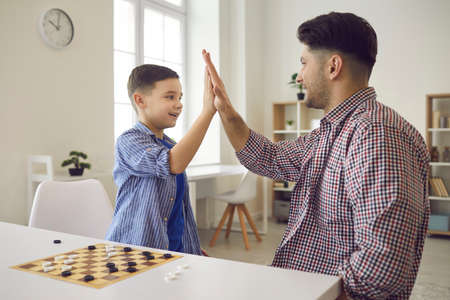 Dad spends time with his son in the game. Joyful little boy gives five to his dad while playing chess at home. Father teaches his son to play logic games such as chess.の写真素材