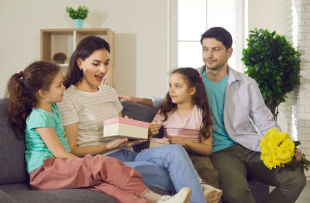 Amazed woman getting congratulations and presents from kids. Loving husband, kid prepare for mum wife gift at birthday or mother day holiday. Happy family sitting on couch in cozy home living roomの写真素材