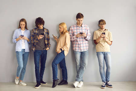 Group of young people standing leaning on wall, using cell phones, chatting online, scrolling...
