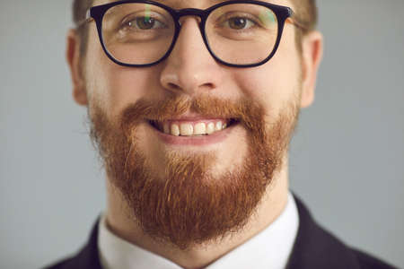 Happy intelligent redhead business man in glasses with ginger beard and mustache and healthy natural straight teeth smiling on gray studio background. Headshot, face closeup shot. Mens style conceptの写真素材