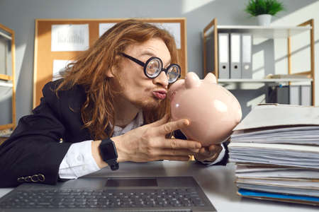 Funny weird looking thrifty man in hilarious round thick lens glasses sitting at office desk with laptop computer and kissing piggy bank. Concept of loving money and saving upの写真素材