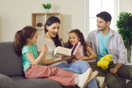 Pleasant surprised mom with happy expression opening gift box from smiling little girl kids daughters and husband. Happy excited family, birthday or mother day holiday celebration togetherの写真素材