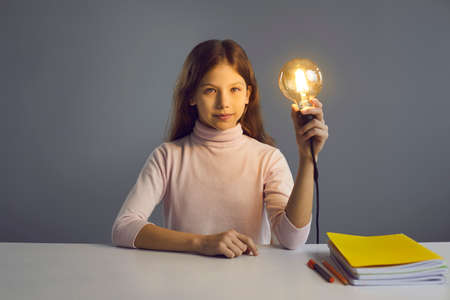 Smart child sitting at desk and holding glowing lit lightbulb. Portrait of serious school girl with shining light bulb at table with books. Creative idea, education, bright student, clever kid conceptの写真素材
