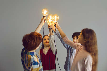 Team of children join glowing lightbulbs. Studio shot of intelligent little kids holding light bulbs. Sharing clever idea, support, collaboration, partnership, making difference, connection, teamworkの写真素材