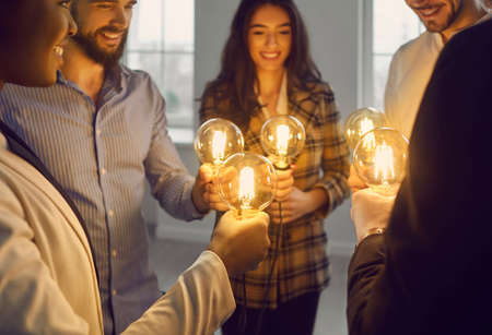 Multiethnic business team standing in circle holding glowing lit retro light bulbs. Group of happy multiracial people join shining vintage Edison lightbulbs. Teamwork, sharing creative ideas conceptの写真素材