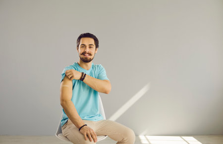 Portrait of happy young man after  shot. Cheerful guy sitting on chair shows arm after flu, coronavirus, tetanus, hepatitis, meningitis or typhoid fever vaccine. Vaccination and safety conceptの写真素材