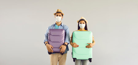 Couple going on a holiday trip. Man and woman in face masks holding suitcases standing isolated on light gray copy space banner background. Concept of people ready for summer vacation and safe travelの写真素材