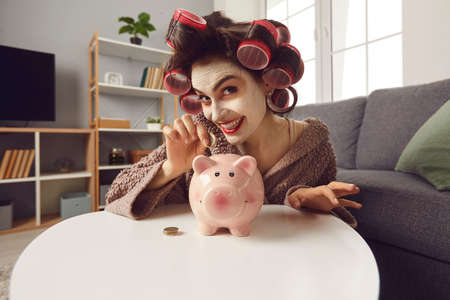 Funny happy young woman, single lady or housewife, with face mask and hair rollers putting coins in pink piggy bank and smiling. Saving up, making plans, family budget management, loving money conceptの写真素材