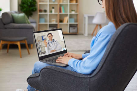 Over shoulder view of young patient sitting in chair communicating with online doctor using laptop. Video telemedicine service, social distancing, Covid 19 pandemic, virtual hospital or clinic conceptの写真素材