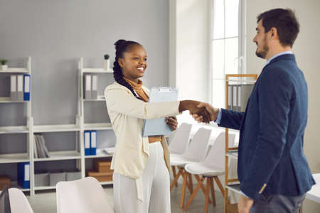 Friendly African american female bank manager shakes hands welcoming a client who has come for legal advice. Man came to the bank office to get advice on his finances, loans, mortgages or insurance.の写真素材