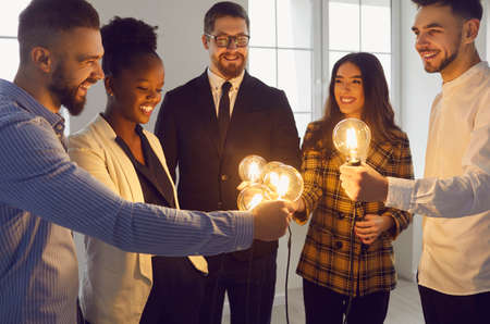 Group of multiracial business people standing in circle and holding glowing lit light bulbs. Team of happy multiethnic entrepreneurs join shining lightbulbs. Teamwork, sharing creative ideas conceptの写真素材