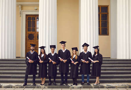 Group of young smiling people university graduates in traditional black bonets and masters mantles standing with dimpomas in hands and looking at camera over university building backgroundの写真素材