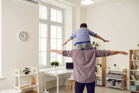 Fathers day. Little boy sits on his dads shoulders and they have fun imitating a flight. Father at home plays with his son by placing him on his shoulder and standing with his back to the camera.の写真素材
