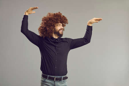 Lets have a party. Profile view of happy young man with crazy curly hair having fun in studio. Side view of carefree guy dancing and doing funny arm moves isolated on gray backgroundの写真素材