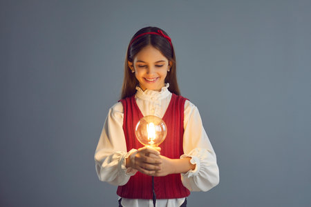 Primary school girl looking at glowing light bulb in hands. Elementary student holding shining electric lightbulb. Child developing own creative idea. Happy kid enjoys learning and making discoveriesの写真素材