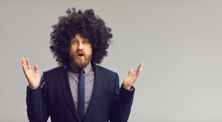Funny young business man in big curly wig gasps in surprise standing on studio copy space background. Astonished guy in office suit with amazing hair looking at camera isolated in gray backgroundの写真素材