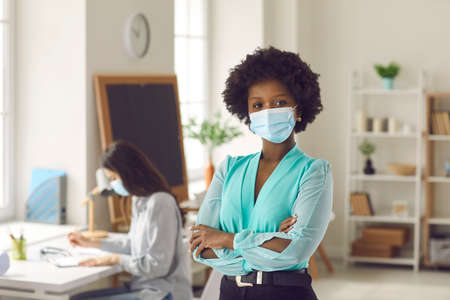 Portrait of black businesswoman with Afro hairstyle in medical face mask standing arms crossed in office. Young woman returns to work after Covid-19 lockdown ends. Coronavirus pandemic safety conceptの写真素材