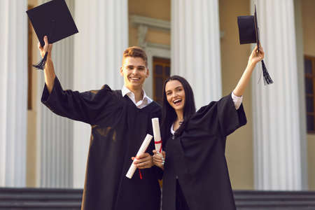 Happy smiling couple girl and boy university graduates standing with dimplmas and holding bonets in raised hands over university building at background. Successful graduating from universityの写真素材