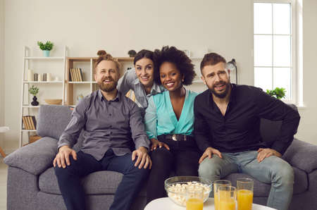 Group of multi racial sports fans sitting on sofa at home and enjoying football game on television. Multiethnic male and female friends supporting different teams while watching soccer match on TVの写真素材
