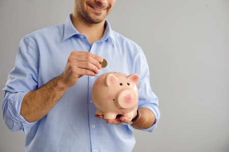 Cropped image of an unknown man standing on a gray background puts a coin in a piggy bank in the form of a pig. Concept of investment, deposits and finance, wealth and accumulation.の写真素材