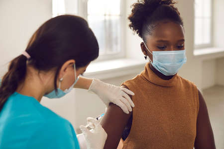 Young black african american woman patient in protective face mask sitting getting vaccination injection against coronavirus from female nurse in clinic.の写真素材