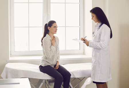 Visit to hospital. Patient telling nurse or clinician about seasonal cold and flu infection symptoms. Young woman complaining about cough and sore throat sitting on examination bed at doctors officeの写真素材
