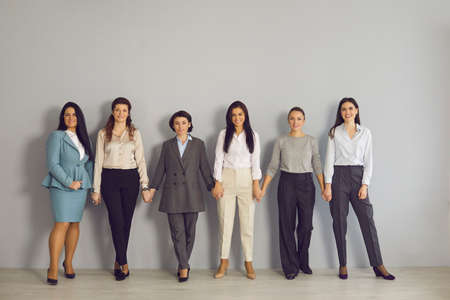 Women business team, cooperation, working in office. Team of young positive businesswomen corporate partners employees standing and holding hands together looking at camera over grey wall backgroundの写真素材