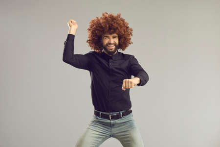 Lets have a party. Happy young man with funny crazy hair having fun in studio. Carefree goofy guy wearing curly wig smiling and dancing  style isolated on gray backgroundの写真素材