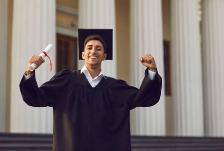 Graduating from university or college. Young happy university graduate standing with diploma in hand and expressing happiness with raised hands over university building backgroundの写真素材