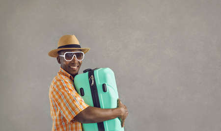 Banner with portrait of happy black tourist with his baggage. African man in sunglasses, sun hat and orange shirt smiling and holding mint green travel suitcase isolated on gray copy space backgroundの写真素材
