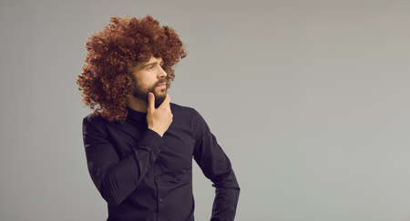 Intrigued man with funny hair looking away standing on grey background with ad copy space. Banner with handsome curious guy in Afro wig rubbing chin thinking over interesting question or wise quoteの写真素材