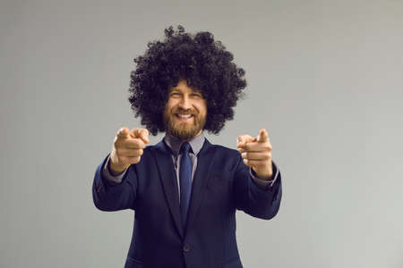 Portrait of funny happy smiling bearded man in formal suit and crazy curly wig pointing double index...