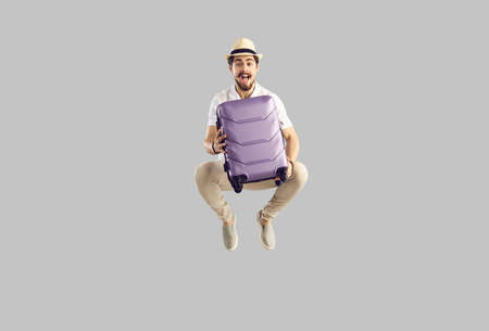 Funny young tourist jumping excited about travelling alone. Portrait of happy overjoyed caucasian man in fedora hat holding purple wheel suitcase and flying in mid air isolated on grey backgroundの写真素材