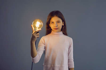 Smart intelligent pre-teen schoolgirl holding glowing idea lamp light bulb studio shot on grey wall background. Rational mind, brainstorming, innovation and solution creation conceptの写真素材