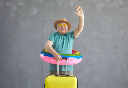 Happy mature tourist leaving for vacation alone. Excited fat senior man in sun hat, pool lifesaver and cool sunglasses holding suitcase and waving goodbye as hes going on travel adventure far awayの写真素材