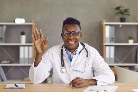 Webcam view. Friendly african american male doctor waving hand greeting patient during video call. Man in a medical gown and glasses sits at his workplace and provides online consultations to patientsの写真素材