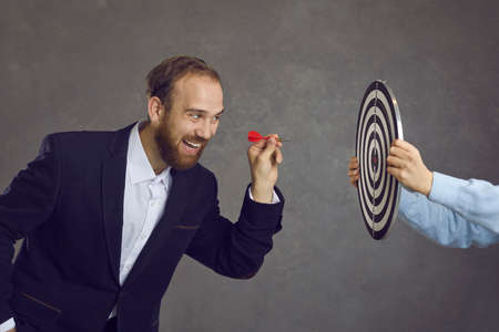 Ambitious businessman aiming red dart at target dartboard determined to throw it and hit bulls eye, side view. Setting business goal, facing challenge, using opportunities, achieving objective conceptの写真素材
