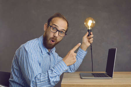 Portrait of funny surprised nerdy bearded man in geek eyeglasses sitting at office desk with laptop computer, looking at camera and showing bright lightbulb hes holding in hand. Amazing idea conceptの写真素材